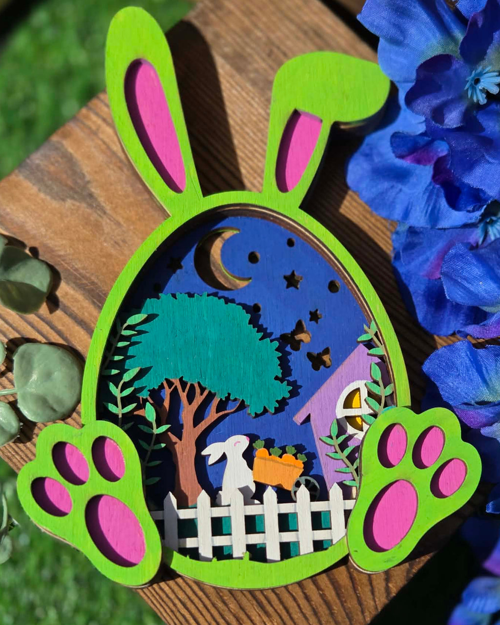 Wooden cutout of a rabbit with a night scene inside, surrounded by blue flowers.